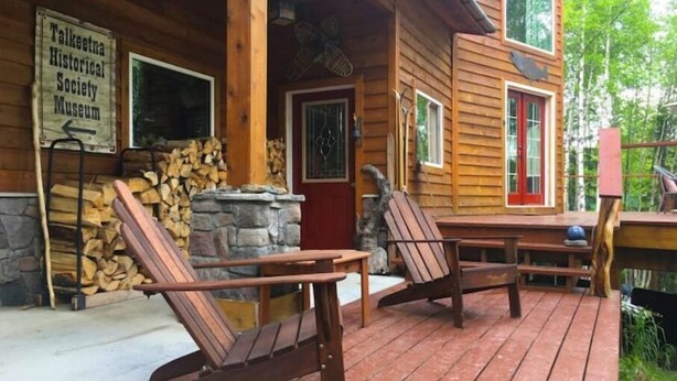 A wooden cabin with a porch, two Adirondack chairs, a stone fireplace, and a sign for the Talkeetna Historical Society Museum. - The Rusty Salmon Lake House with Deck and Dock (Talkeetna)