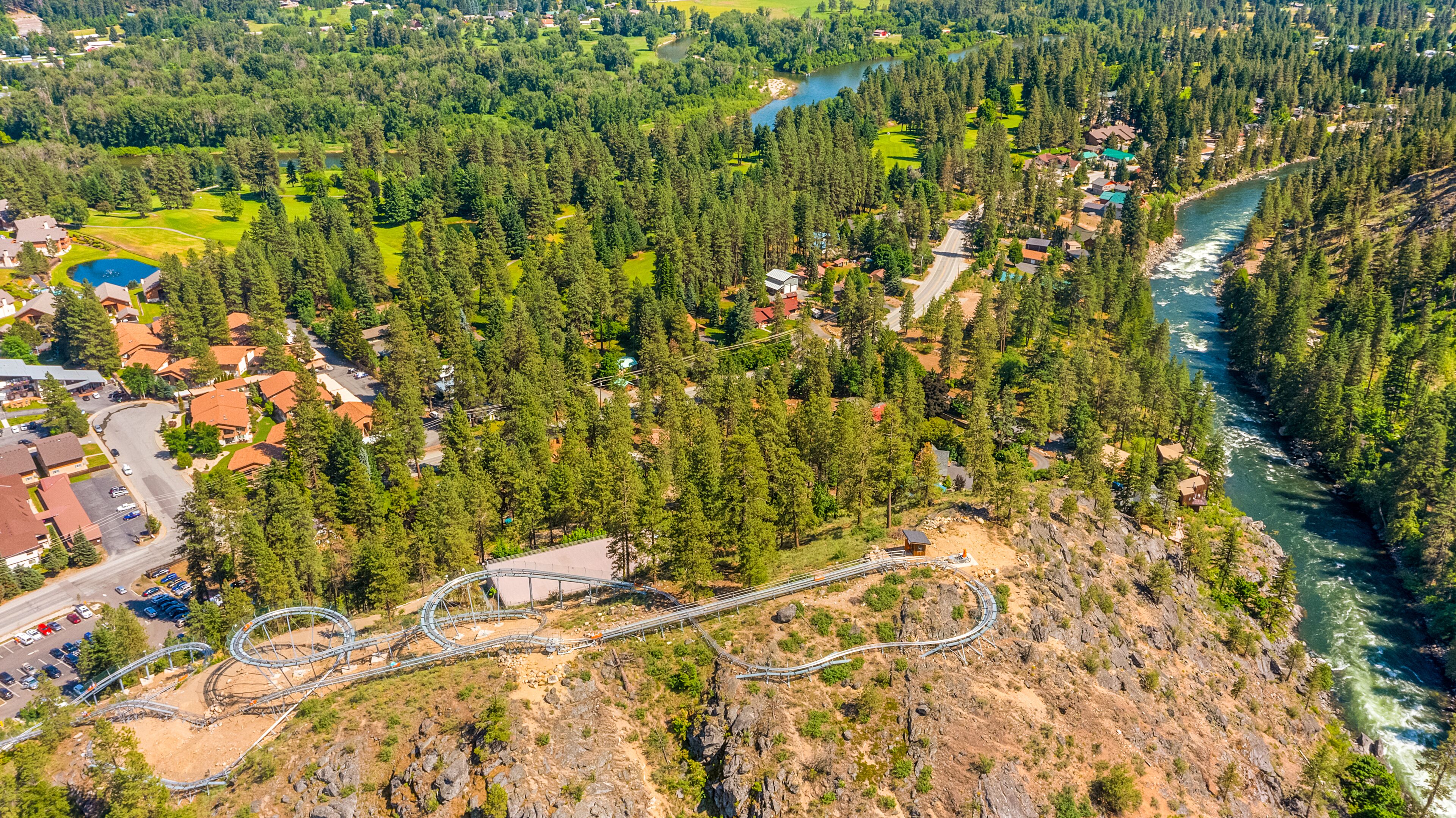 An aerial view of a mountainous area with a winding road, a river, and a small cabin.