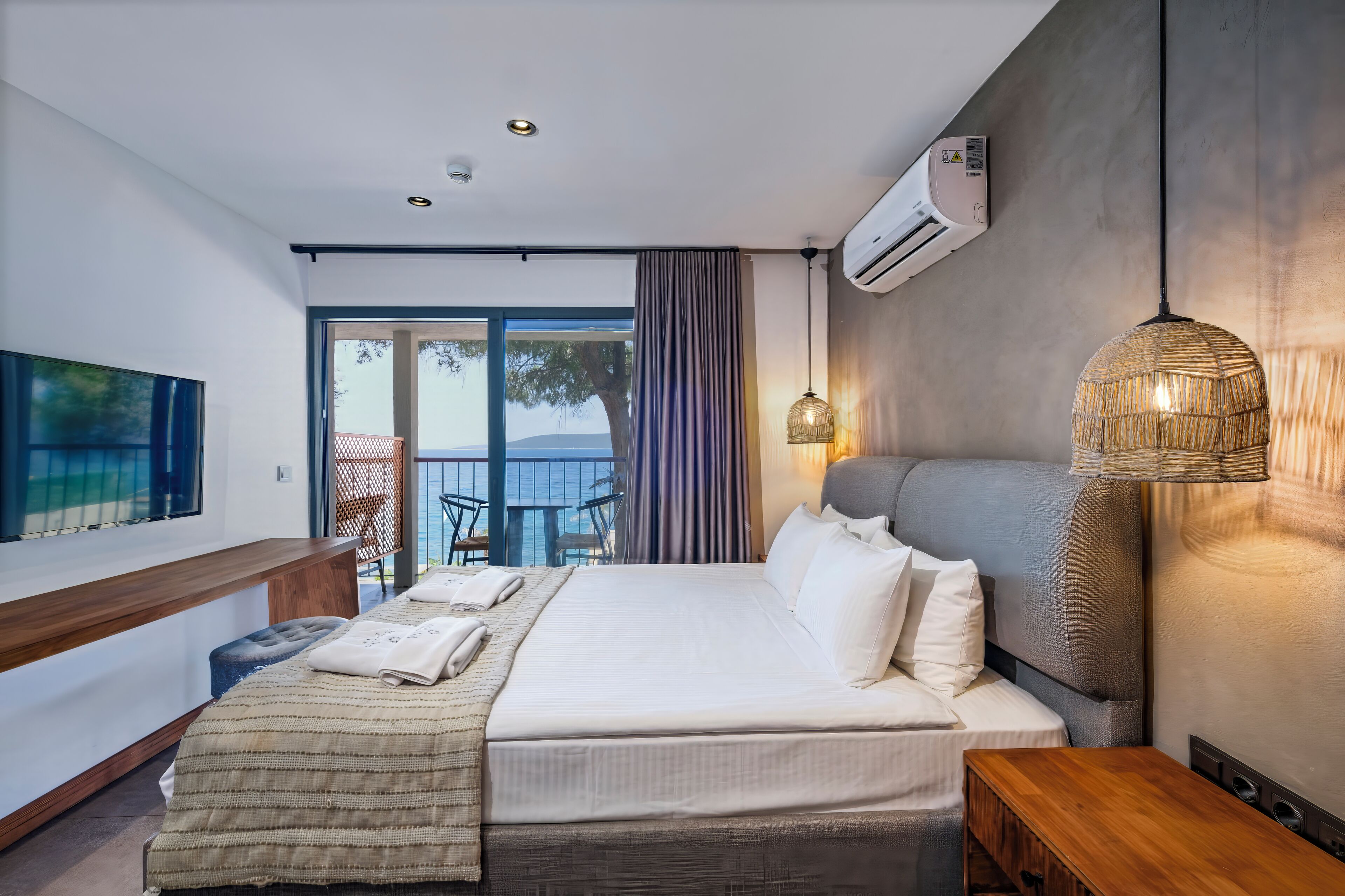 A modern hotel room with a large bed, a wooden bench, a small table, a wall-mounted air conditioner, and a view of the outdoors.