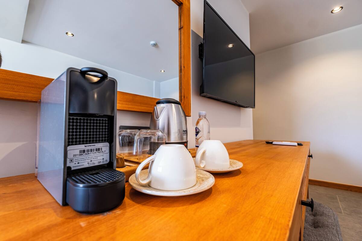 Una cafetera, dos tazas blancas, una botella y un televisor sobre una mesa de madera en una habitación con techo inclinado e iluminación empotrada.