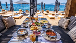A dining table with a variety of dishes and drinks, surrounded by lounge chairs and umbrellas, overlooking the sea. - Isolina Bodrum (Bodrum)