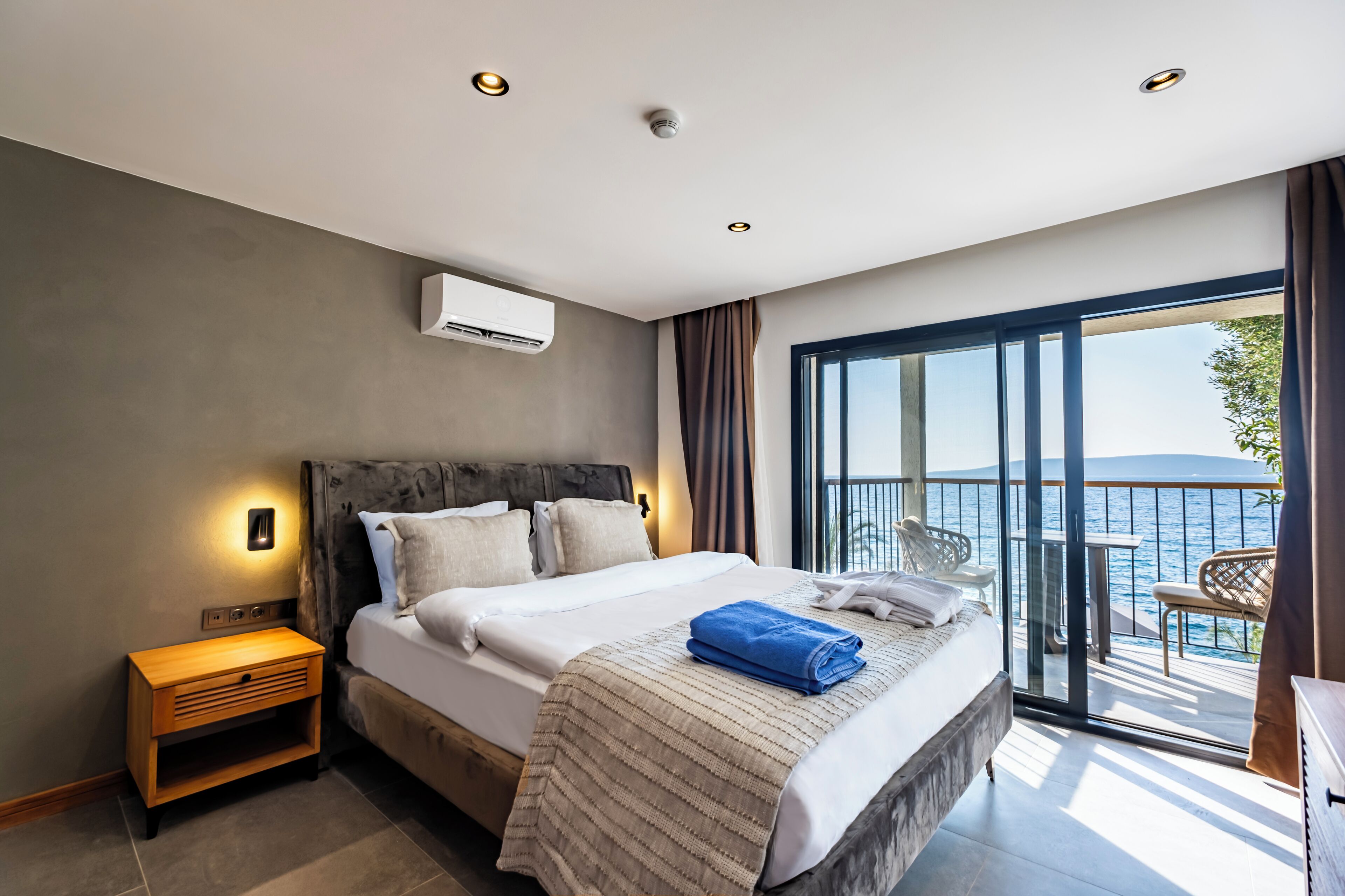 A modern bedroom with a large bed, a nightstand, a window with a view of the sea, and a balcony with outdoor seating.