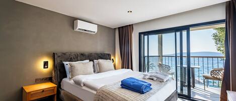 A modern bedroom with a large bed, a nightstand, a window with a view of the sea, and a balcony with outdoor seating.