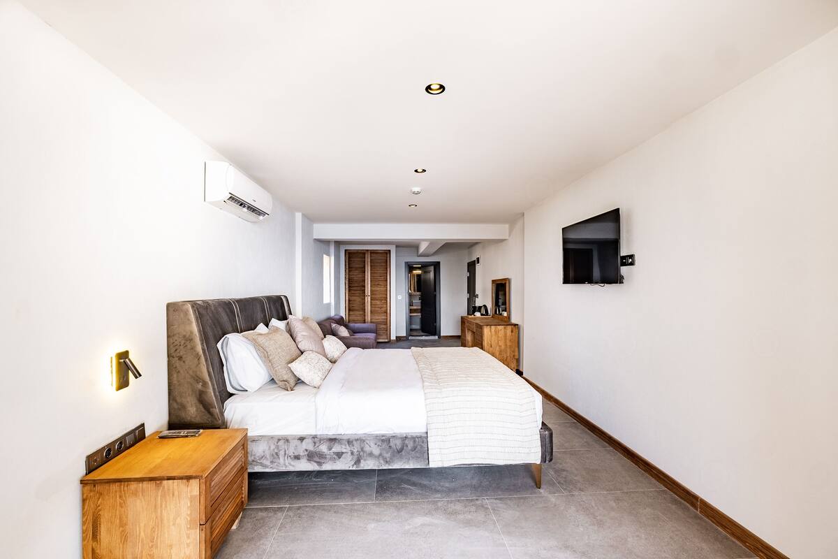 Una habitación de hotel moderna con una cama grande, mesitas de noche de madera, un televisor de pantalla plana y un pasillo que conduce a un baño y otra habitación.