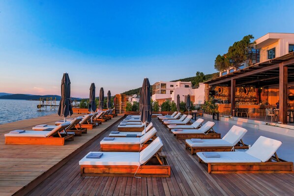 A wooden deck with sun loungers and umbrellas, a bar area, and buildings in the background.