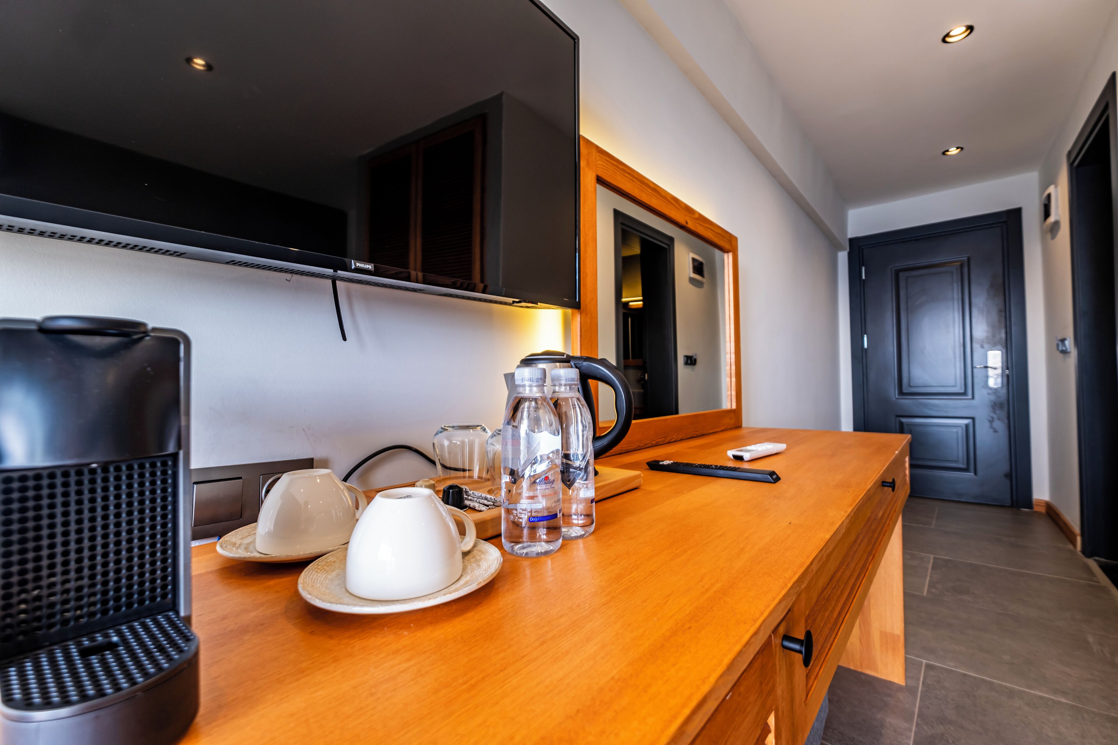 A hotel room with a wooden counter, a coffee maker, a kettle, and a cup on the counter.