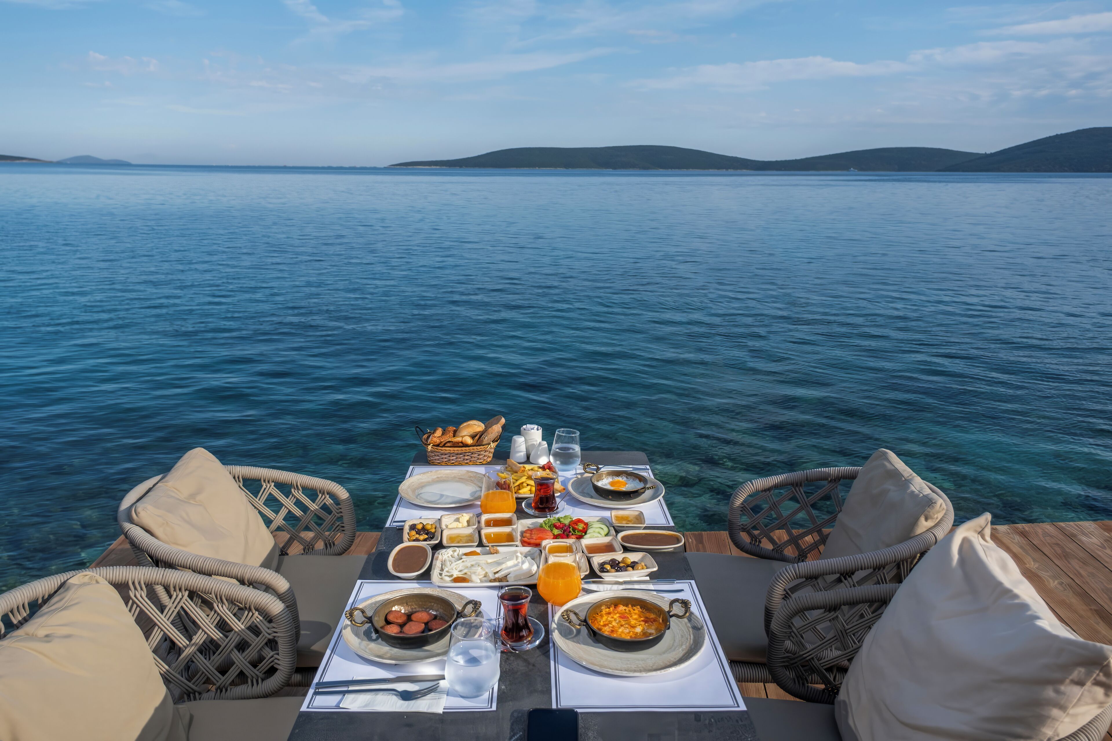 A dining table with various dishes and drinks, overlooking a body of water with hills in the distance.