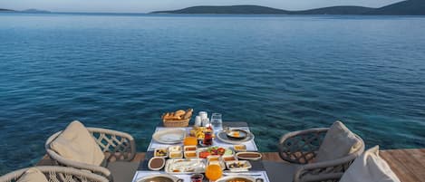 A dining table with various dishes and drinks, overlooking a body of water with hills in the distance.