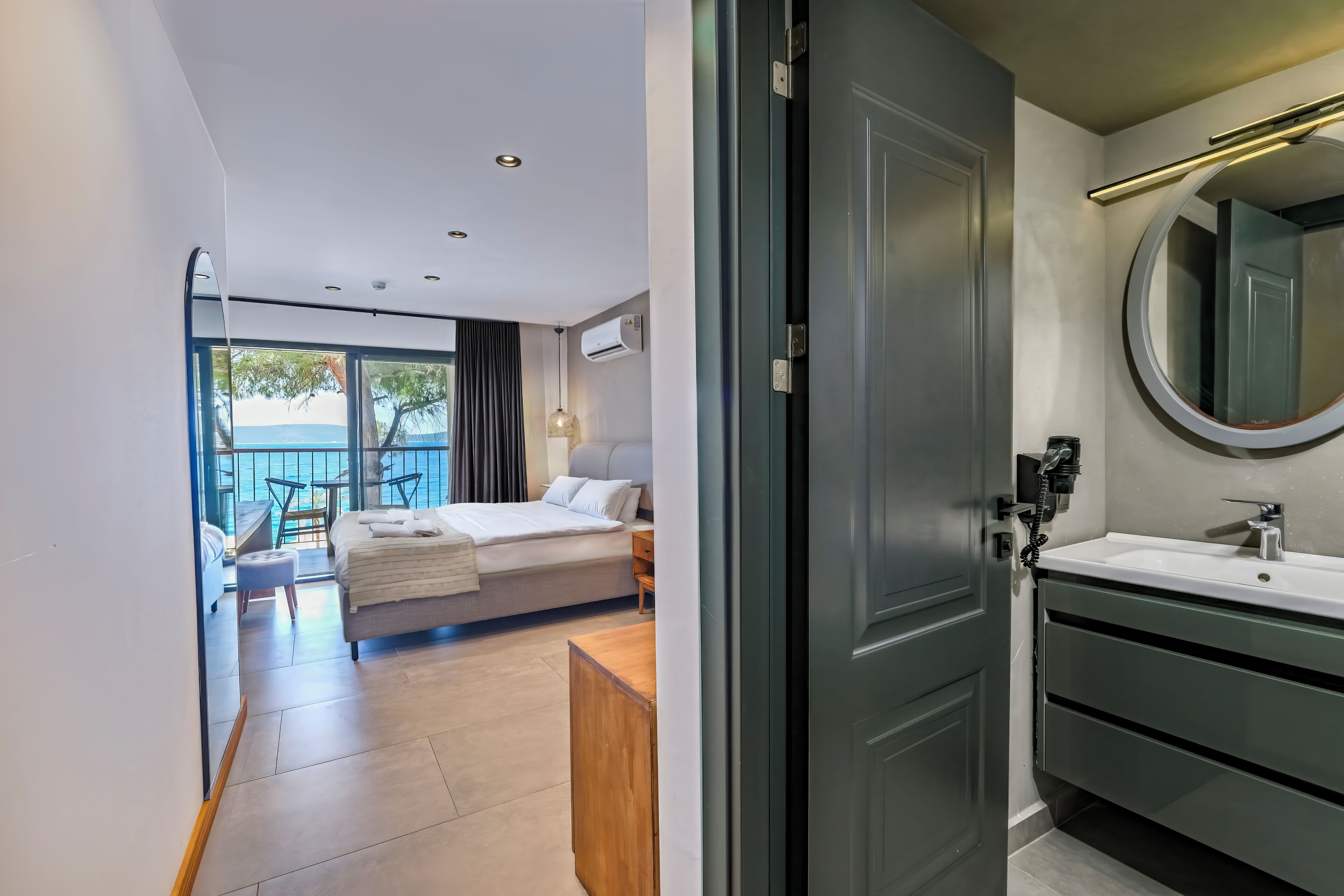 Une chambre d’hôtel moderne, dotée d’un lit, d’une salle de bain avec un miroir et offrant une vue sur la mer grâce à de grandes fenêtres.