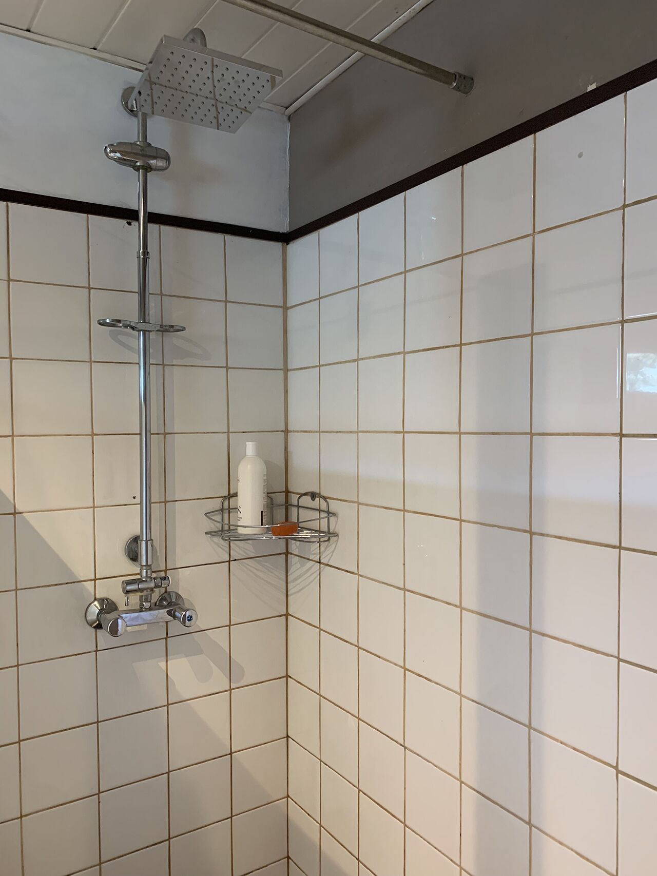 A shower with a handheld showerhead and a wall-mounted faucet. There is a small shelf with toiletries.