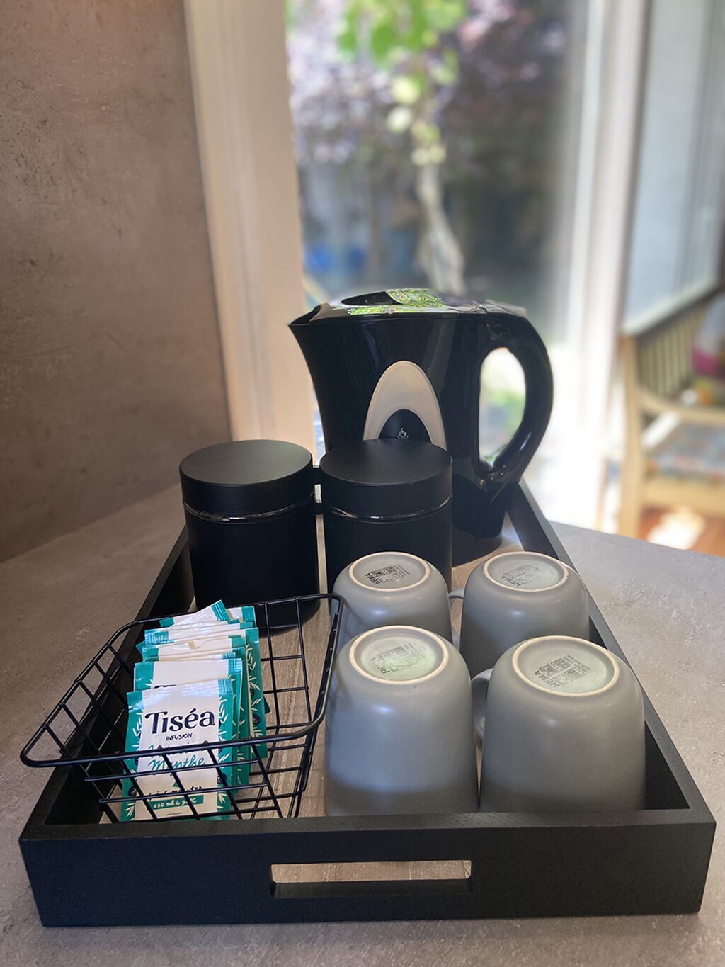 A tray with a black pitcher, two black containers, and four gray bottles, along with a basket of green and white tea packets.