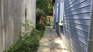 A narrow concrete pathway flanked by a blue house and a grey wall, with a drain grate at the bottom. - Typée, Accueillante et Confortable Maison (Pessac)