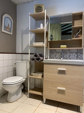Un baño con inodoro, un tocador de madera con cajones, un estante con toallas y un salpicadero de azulejos.