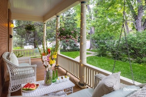 A covered outdoor seating area with wicker furniture, a table with a watermelon and flowers, and a hanging plant. - Pet-Friendly! Stunning Home — Steps to Downtown, CU & Hiking Trails. (Boulder)