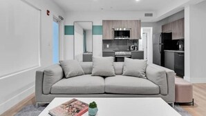 Ein modernes Wohnzimmer mit einem grauen Sofa, einem weißen Couchtisch und einer kleinen Pflanze. Im Hintergrund ist eine Küche mit dunklen Schränken und einem schwarzen Kühlschrank zu sehen.