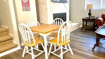 A dining area with a wooden table and white chairs, a stone fireplace, and a television mounted above it.