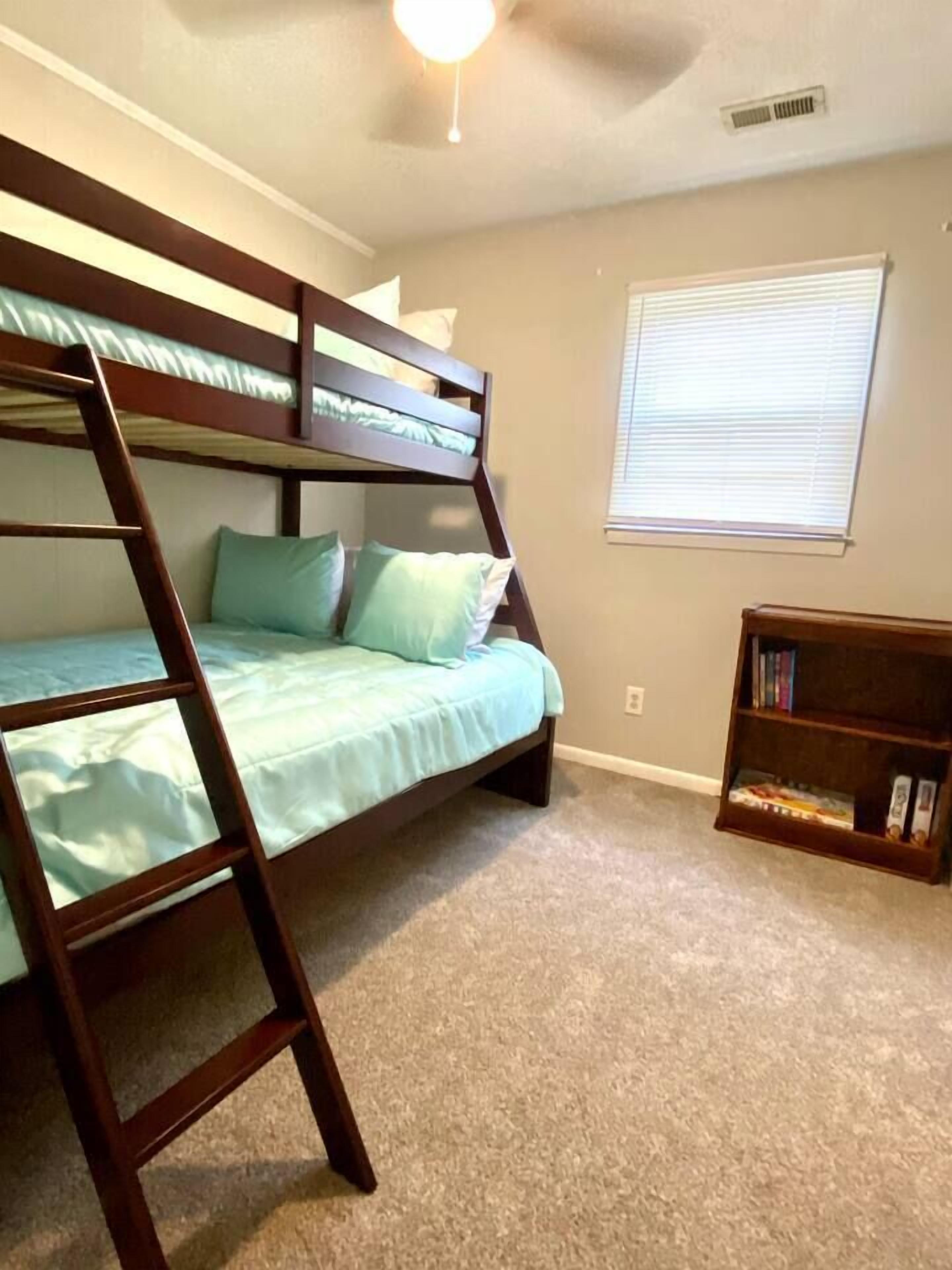 Une chambre avec un lit superposé, un ventilateur de plafond, une bibliothèque et une fenêtre avec des stores.