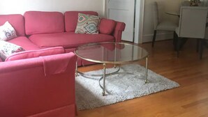 A living room with a red sofa, a glass coffee table, and a painting of a cityscape. - Windsor Lofts 3 (Roanoke)