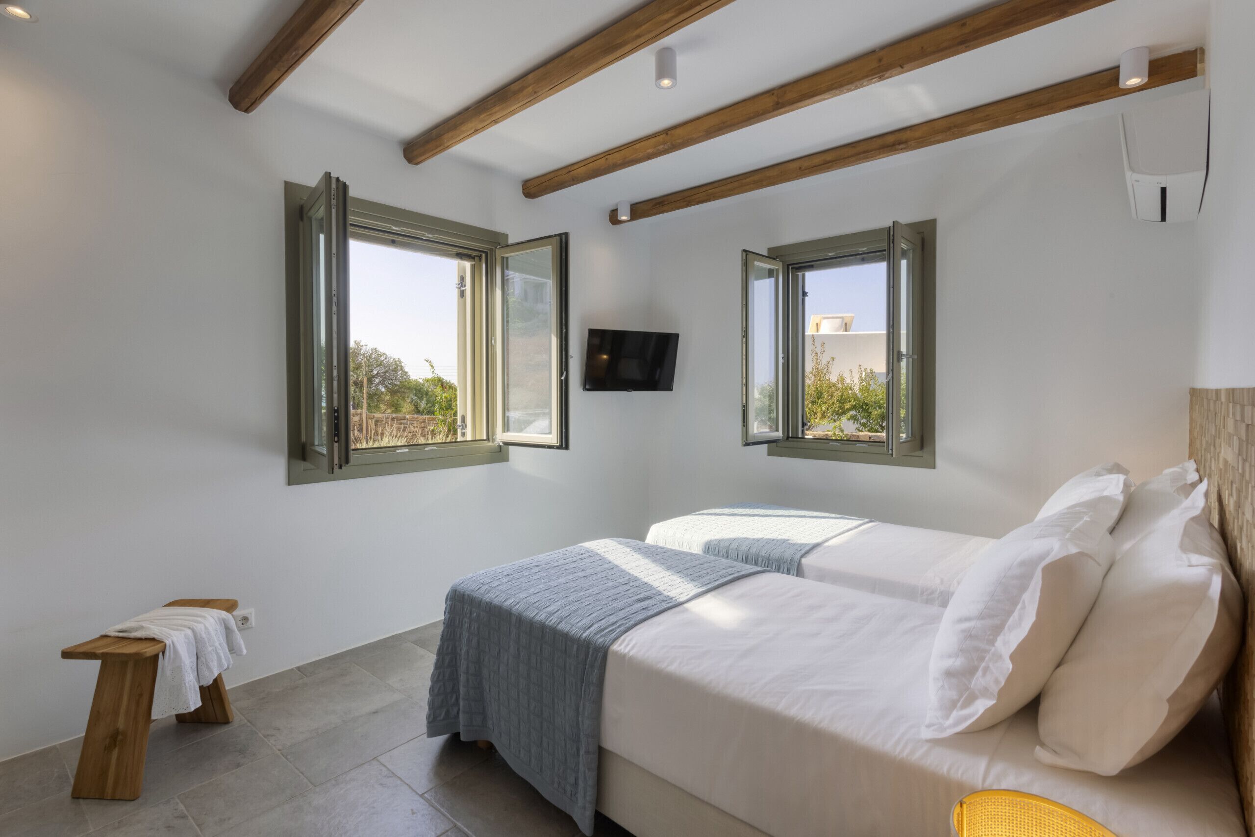 Un dormitorio con dos camas, vigas de madera en el techo, un televisor montado en la pared y ventanas grandes con contraventanas abiertas.