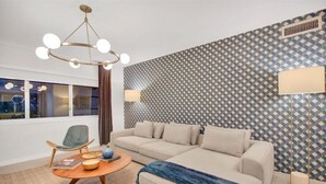 Un salon moderne avec un canapé, un fauteuil et une table basse. Les murs sont recouverts d’un papier peint à motifs et le plafond est agrémenté d’un luminaire.