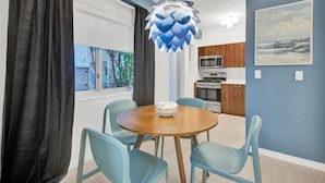 Un coin repas avec une table ronde et quatre chaises, un luminaire suspendu moderne, une cuisine en arrière-plan et un tableau au mur.