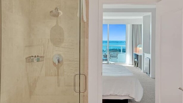 A hotel room with a large window, a bed, a shower area, and a balcony with a view. - Junior Suite 33rd Flr in Fontainebleau Miami Beach (Miami Beach)