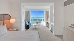A hotel room with a bed, a desk, a chair, a small table, and a balcony with a view of the ocean. - Junior Suite 33rd Flr in Fontainebleau Miami Beach (Miami Beach)