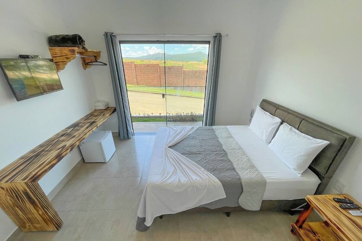 Une chambre à coucher comprenant un lit, un banc, une petite table et une fenêtre donnant sur un mur de briques et de la verdure.
