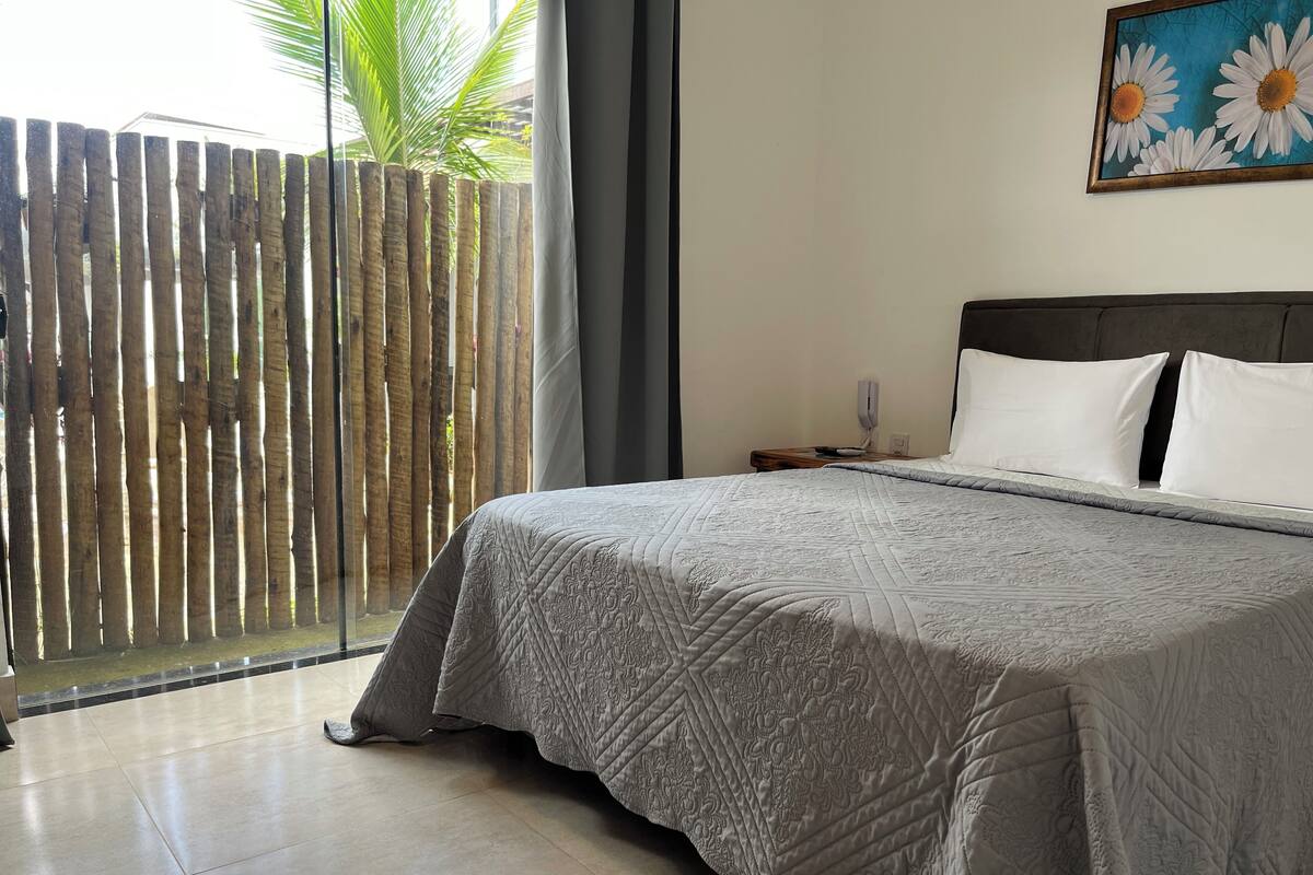 Une chambre avec un grand lit, une table de chevet, un téléphone fixé au mur et une fenêtre donnant sur une clôture en bambou et un palmier.
