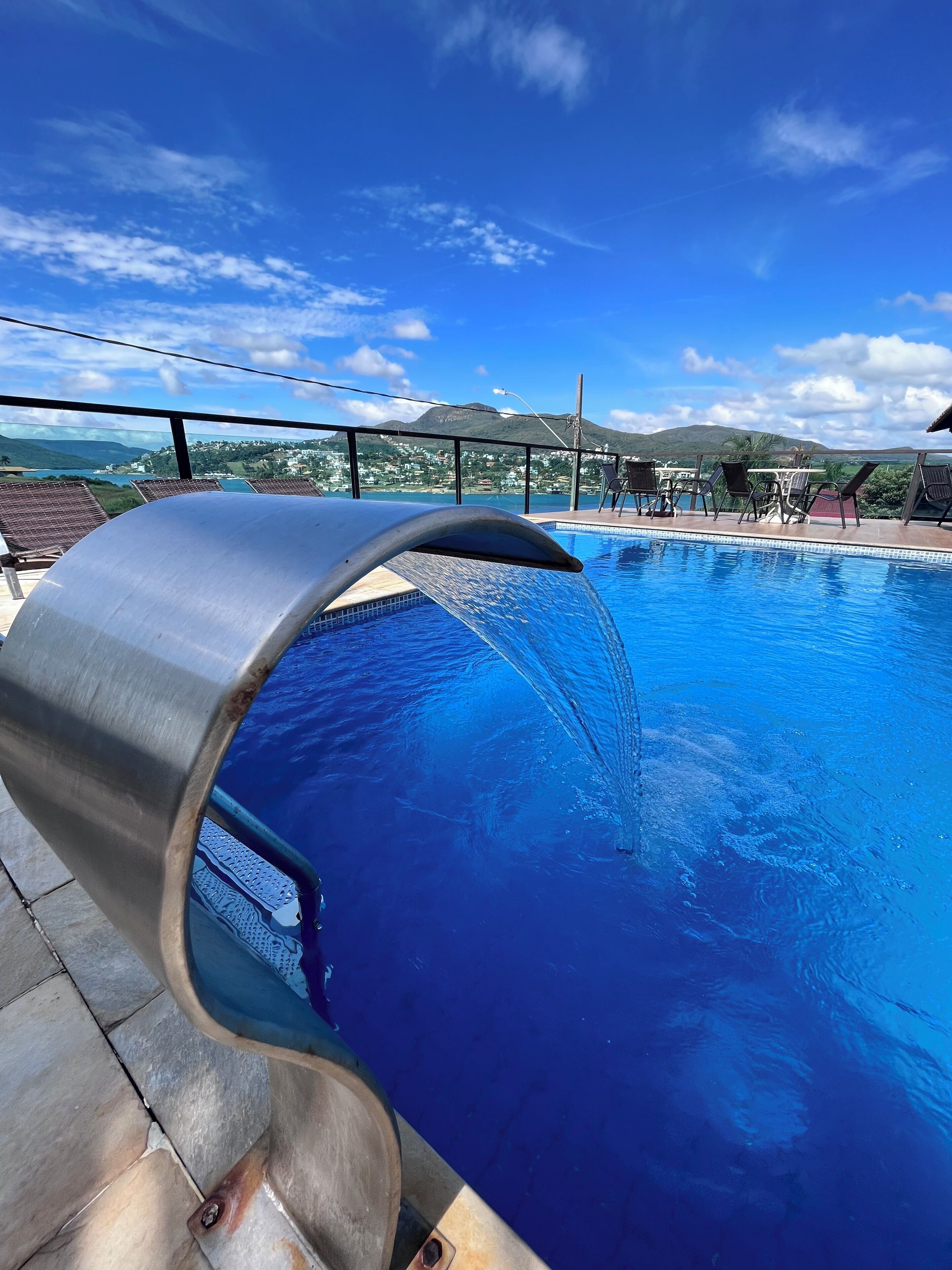 Una piscina con cascada, rodeada de sillas de descanso y una vista panorámica de montañas y un pueblo.