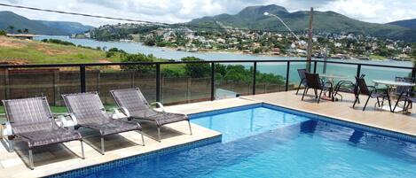 Piscina con sillas de descanso y una mesa, con vistas a un lago y colinas.