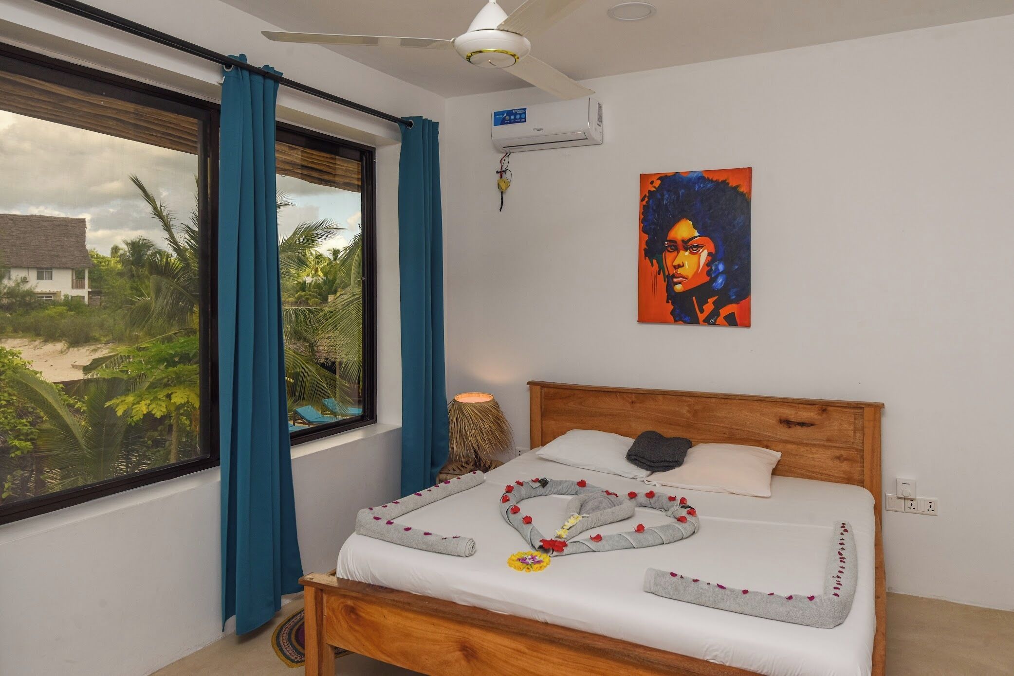 Une chambre dotée d’une grande fenêtre, d’un ventilateur de plafond, d’un climatiseur, d’un lit avec une tête de lit en bois et d’un tableau accroché au mur.