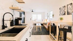 Una cucina moderna con un rubinetto nero, scaffali in legno e un piano di lavoro nero. C'è una zona pranzo con tavolo e sedie, e un divano sullo sfondo.