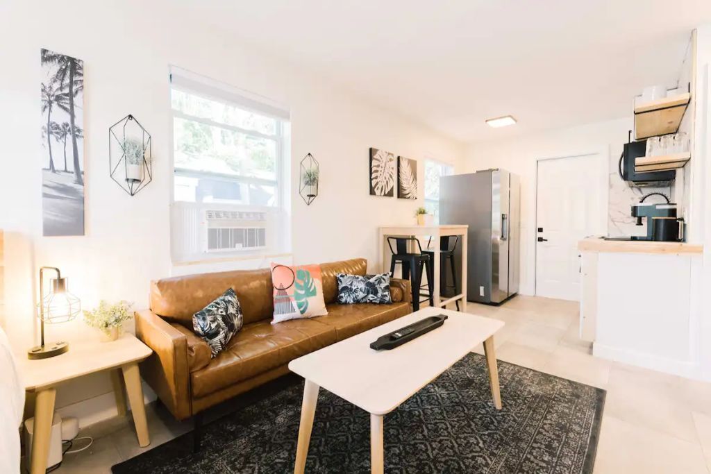 Una sala moderna con un sofá de cuero marrón, una mesa de centro blanca y una cocina con refrigerador y encimera.
