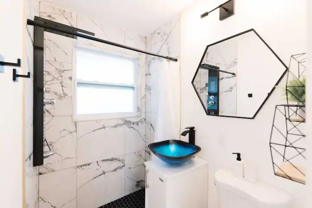 Un baño moderno con un salpicadero de mármol blanco, un lavabo tipo cuenco azul, un lavamanos pedestal blanco, un espejo geométrico y una ducha con puerta negra.