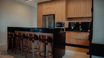 Cozinha moderna com armários de madeira, geladeira de aço inoxidável, micro-ondas e bancada preta. Há três banquetas de madeira com pernas de metal.