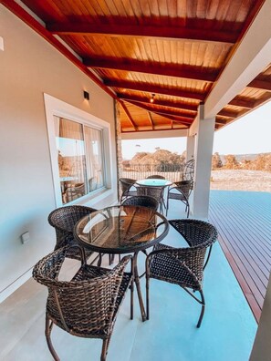 A covered patio with a round glass table and wicker chairs, overlooking a scenic landscape. - Casa Paraíso do Sol - Rancho Queimado (Rancho Queimado)