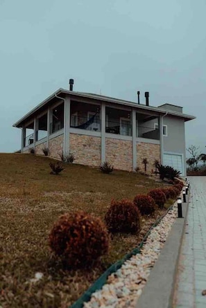 A modern two-story house with a stone facade, a covered porch, and a small garden with shrubs. - Casa Paraíso do Sol - Rancho Queimado (Rancho Queimado)