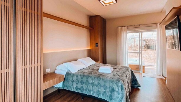 A modern bedroom with a wooden floor, a bed with a blue quilt, a bedside table, and a large window with curtains.