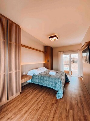 A modern bedroom with a wooden floor, a bed with a blue quilt, a bedside table, and a large window with curtains. - Casa Paraíso do Sol - Rancho Queimado (Rancho Queimado)