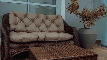 Un canapé en osier doté d’un dossier capitonné et accompagné d’un pouf assorti, placé devant une grande fenêtre offrant une vue sur des plantes de bambou.