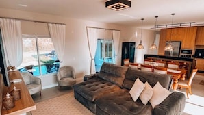 A modern living room with a large sectional sofa, a dining area, and a kitchen with wooden cabinets and pendant lighting.