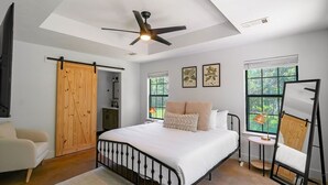 A bedroom with a bed, a ceiling fan, a wooden wardrobe, a chair, a small table, and a window with a view of greenery.