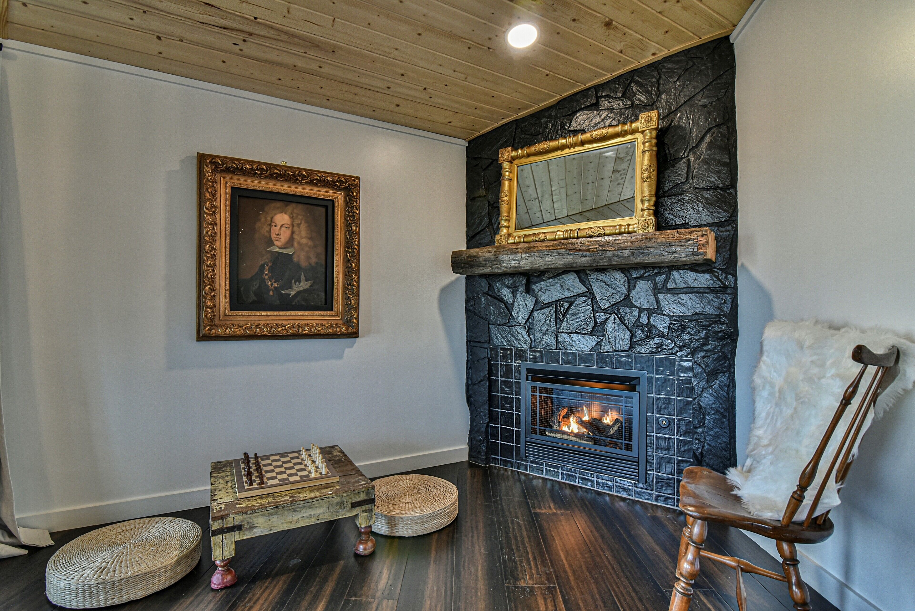 A cozy room with a fireplace, a chessboard, a framed portrait, and a comfortable chair.