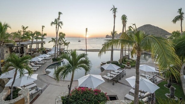 A resort pool area with lounge chairs and umbrellas, overlooking the sea and a mountain at sunset.