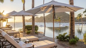 A poolside area with lounge chairs and umbrellas, palm trees, and a view of the sea and mountains.