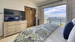 A bedroom with a large bed, a TV on a dresser, a view of the sea, and a balcony.
