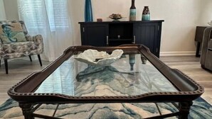 Una mesa de centro con tapa de vidrio y estructura oscura, una alfombra estampada, un mueble de madera oscura y una silla con cojín.