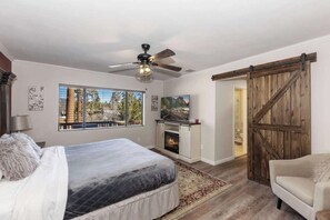 A bedroom with a bed, a ceiling fan, a fireplace, a large window with a view, and a sliding barn door. - Walk to Lake! 7bdr, Hottub, Movie Theater, Gameroom Dawn and Dusk by Avantstay (Big Bear Lake)