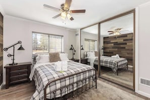 A bedroom with a bed, bedside tables, a ceiling fan, a large mirror, and a window with blinds. - Walk to Lake! 7bdr, Hottub, Movie Theater, Gameroom Dawn and Dusk by Avantstay (Big Bear Lake)